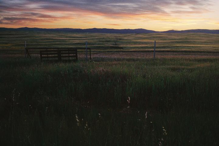 A wooden fence and posts stand in a grassy field with mountains in the distance under a colorful sunset sky.