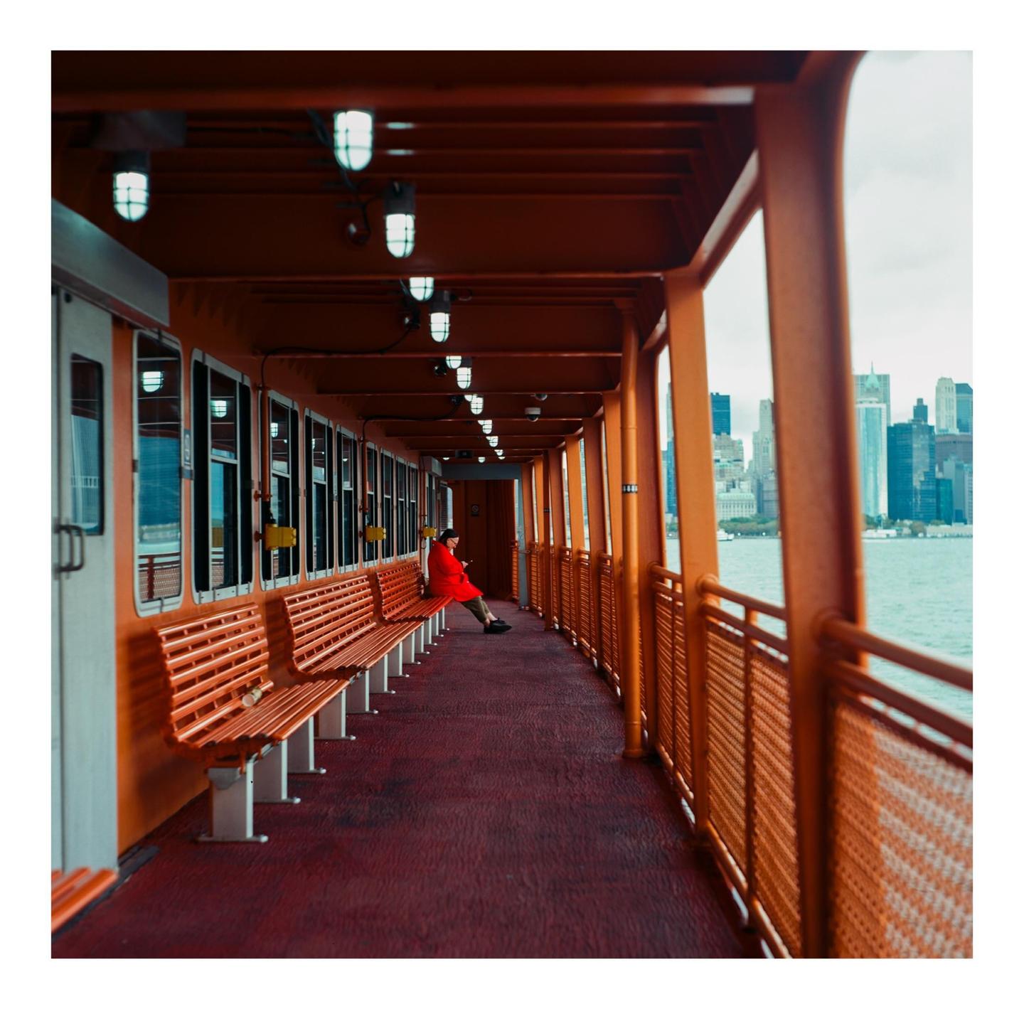 A woman in a red coat sits on a bench on a ferry with a city skyline in the background.