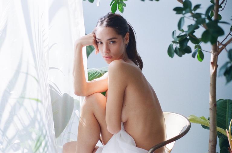 A woman sits on a chair by a window with plants and sheer curtains.