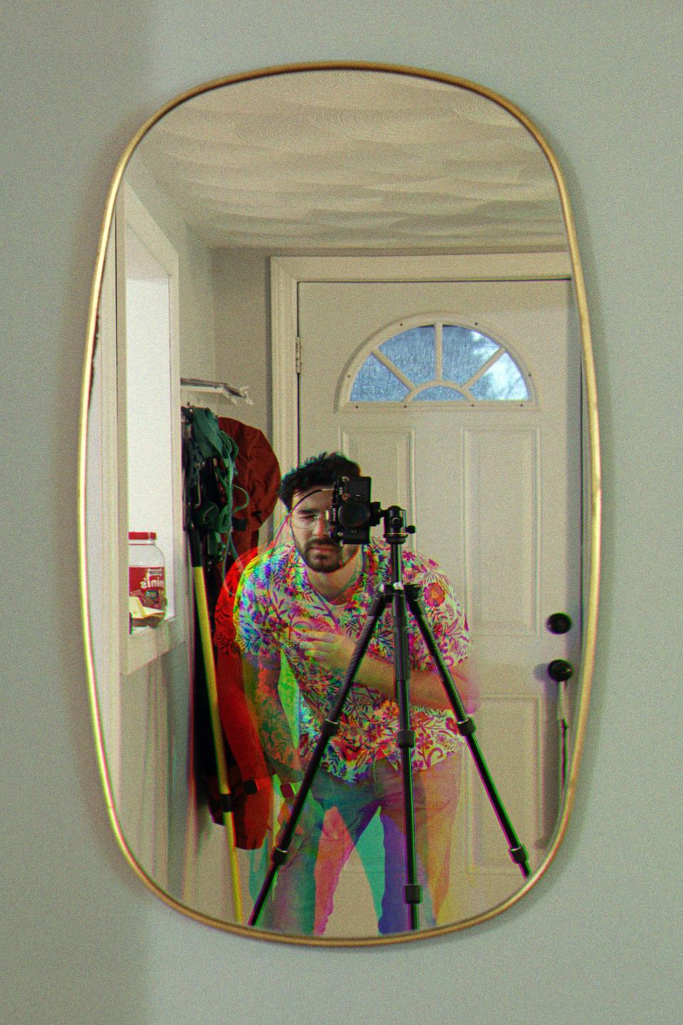 A man in a colorful shirt is reflected in a mirror, holding a camera on a tripod.