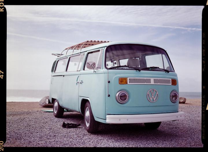 A light blue Volkswagen van is parked on a gravel beach with the ocean in the background.