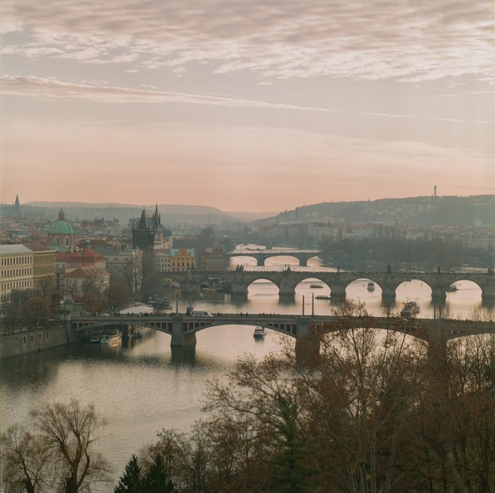 A panoramic view of Prague shows multiple bridges crossing a river, with historic buildings and distant hills under a cloudy sky.
