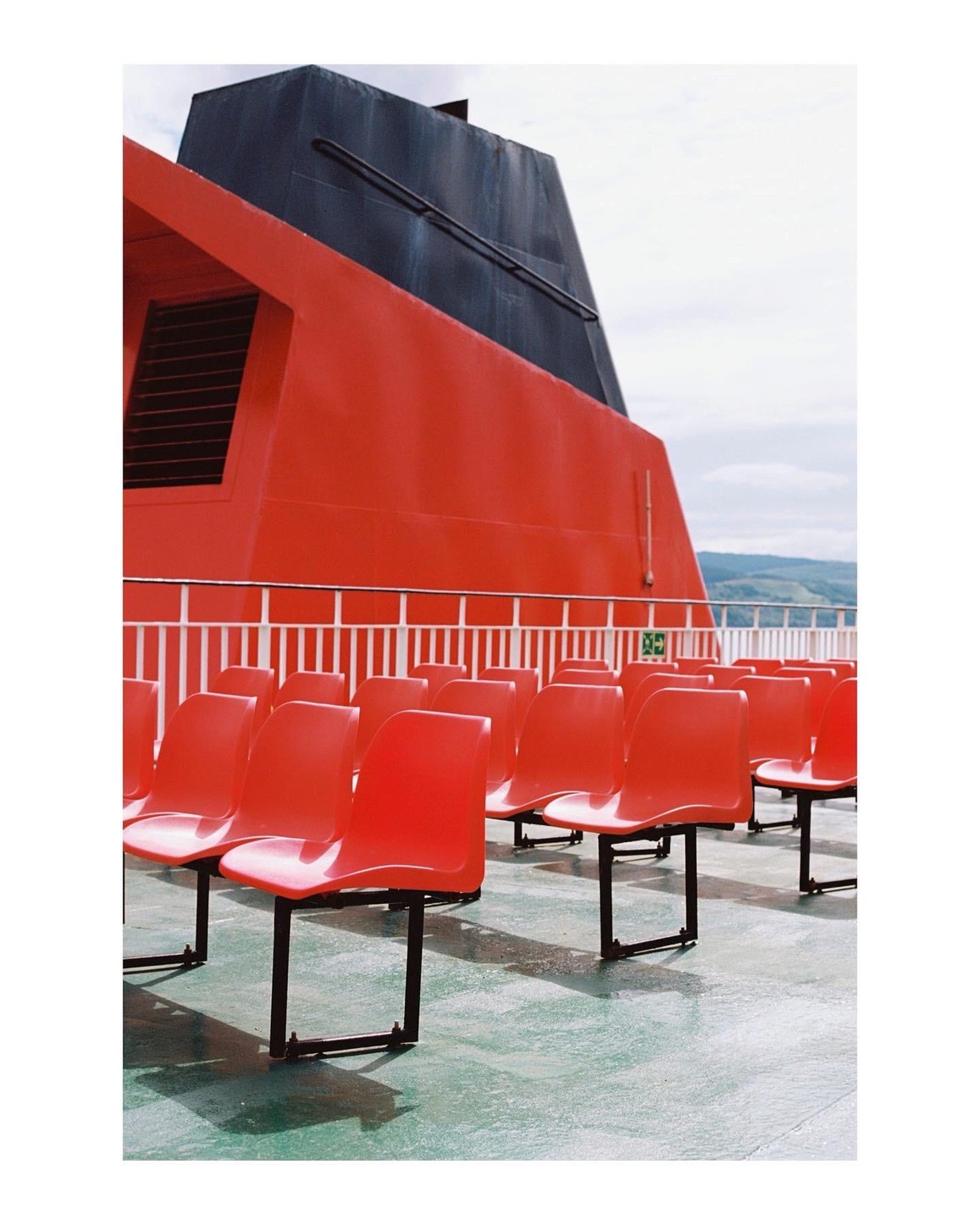 Rows of empty red seats are on the deck of a ferry with a large red and black structure in the background.