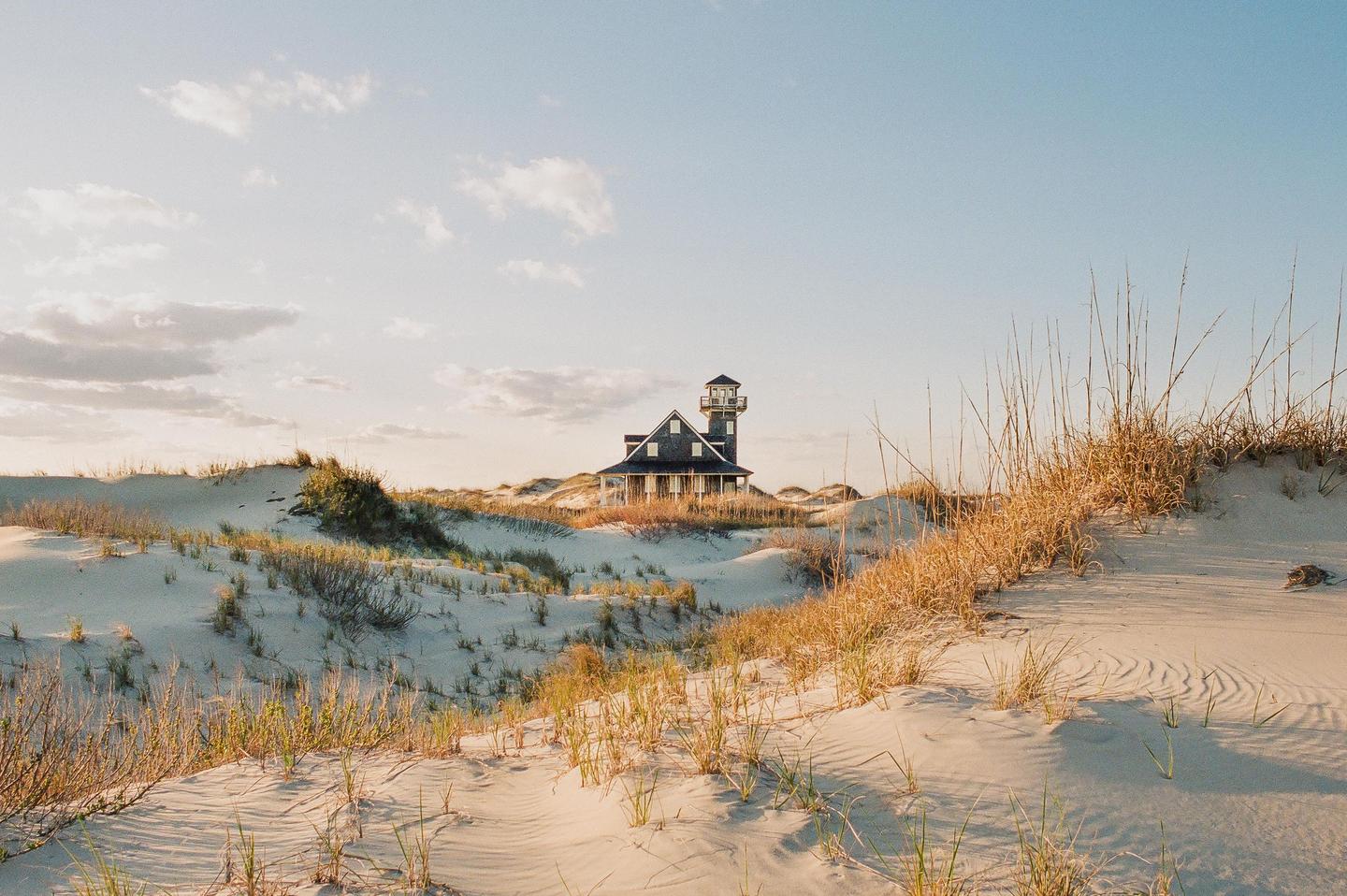 A house with a tower stands among sand dunes covered in dry grass under a partly cloudy sky.