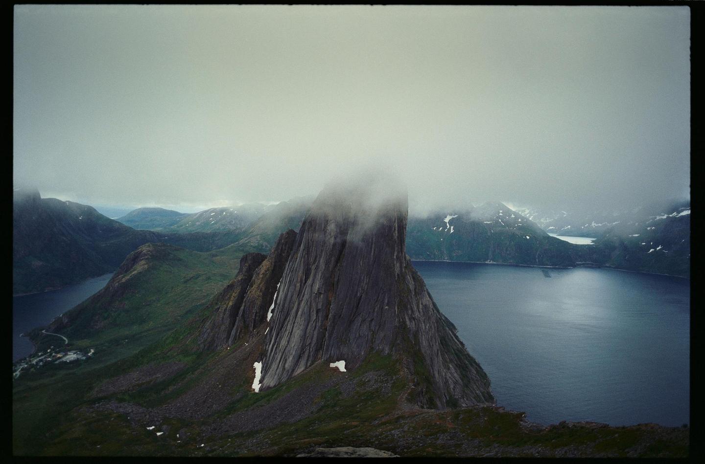 A tall, jagged mountain peak rises from a body of water, surrounded by other mountains under a cloudy sky.