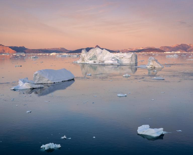 Icebergs float on the water with mountains in the background under a pink sky.