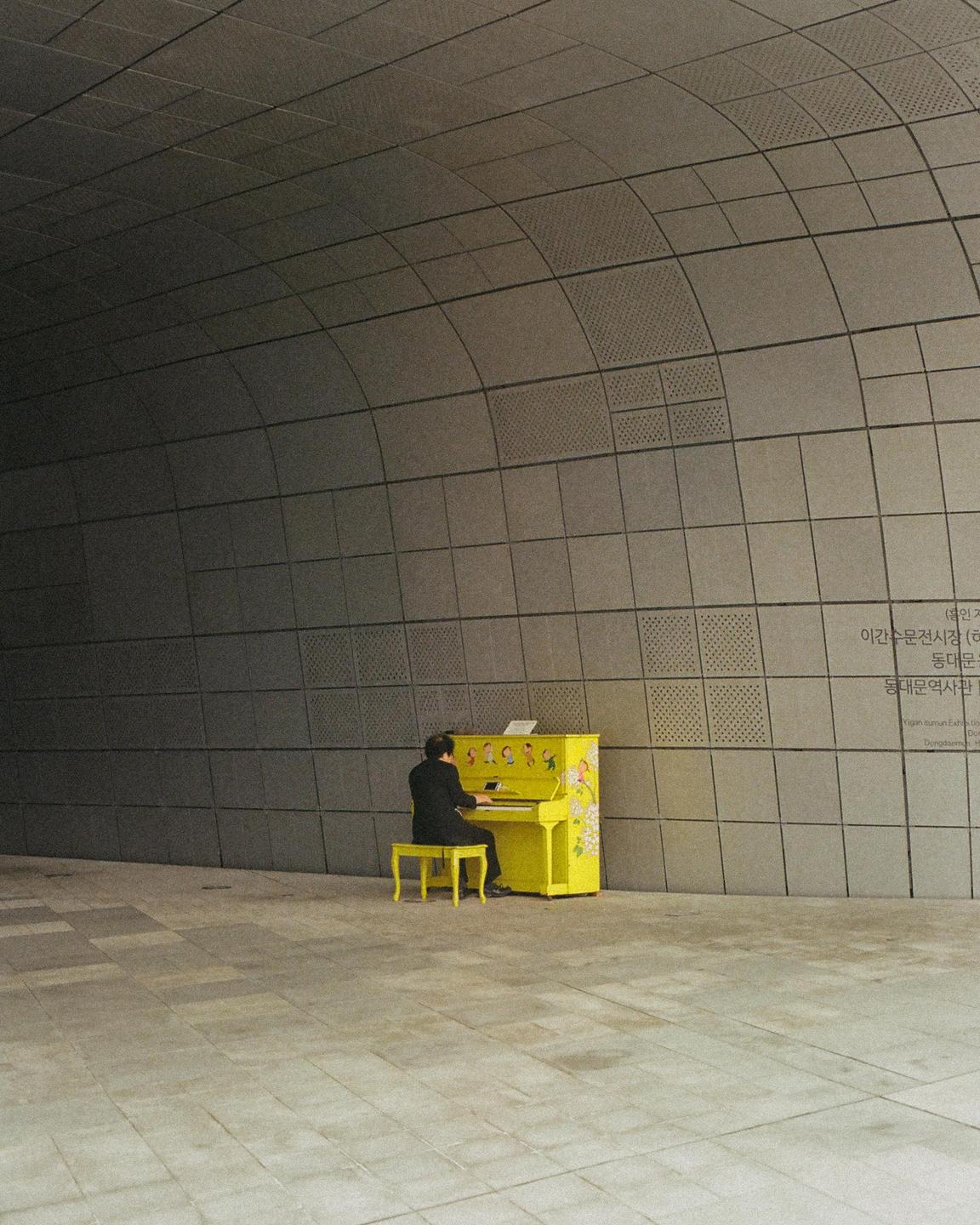 A man plays a bright yellow piano with floral designs in a modern, curved building.