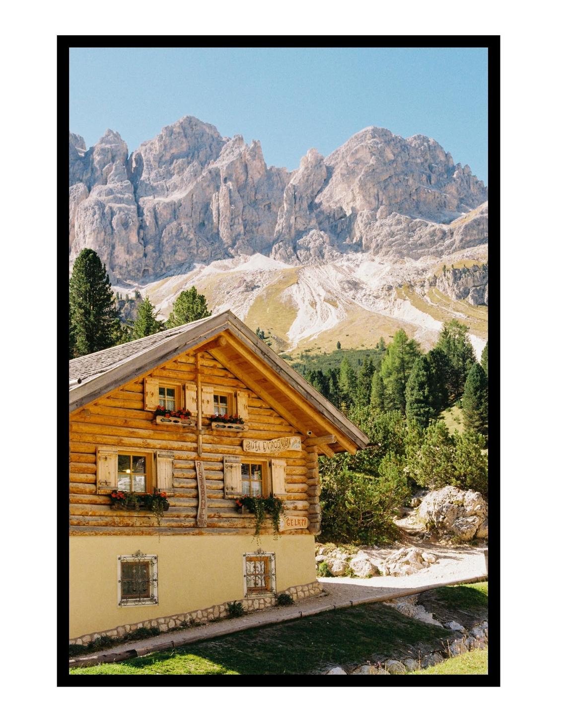 A wooden cabin with flower boxes sits in front of a mountain range with pine trees.