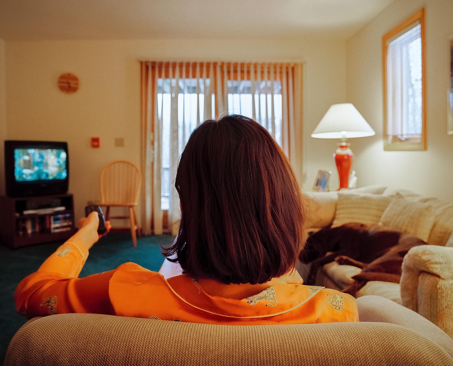A woman with short brown hair sits on a sofa, holding a remote control and watching television.