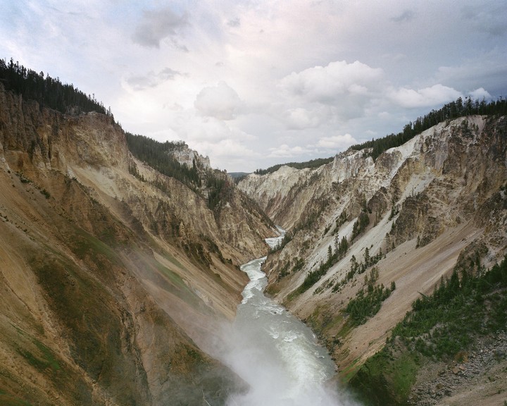 A river flows through a deep canyon with steep, forested slopes under a cloudy sky.