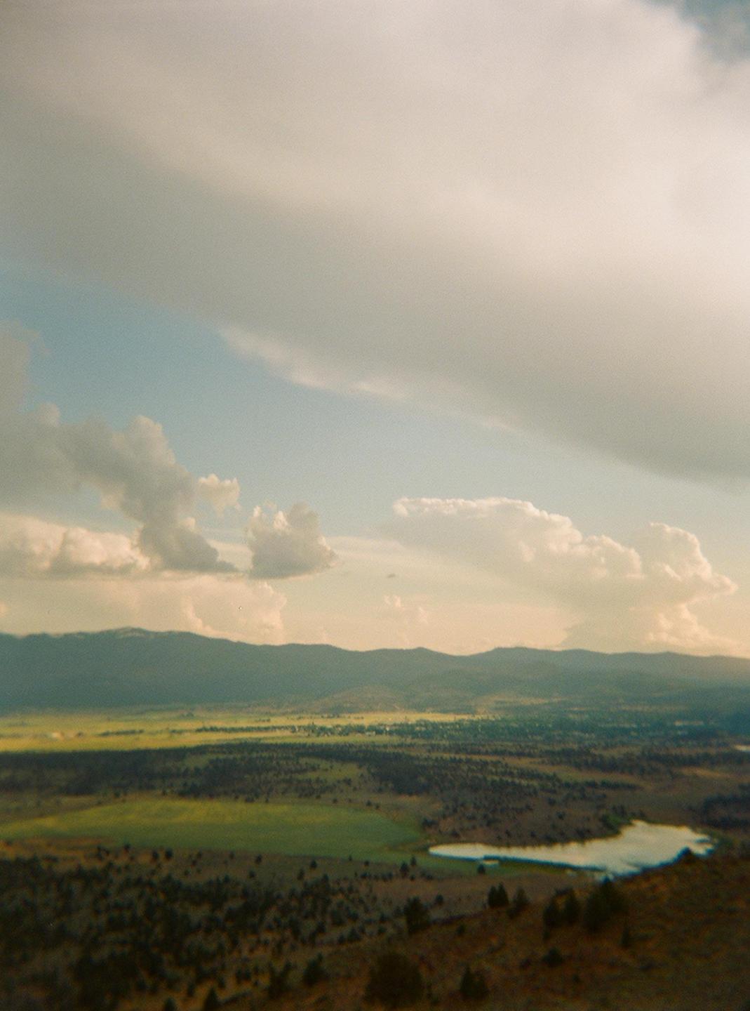 A wide landscape shows a valley with a lake, fields, and mountains under a cloudy sky.