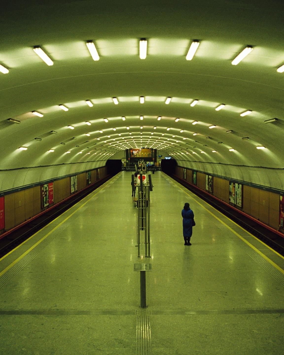 A person stands on a subway platform with a long, arched ceiling illuminated by many fluorescent lights.