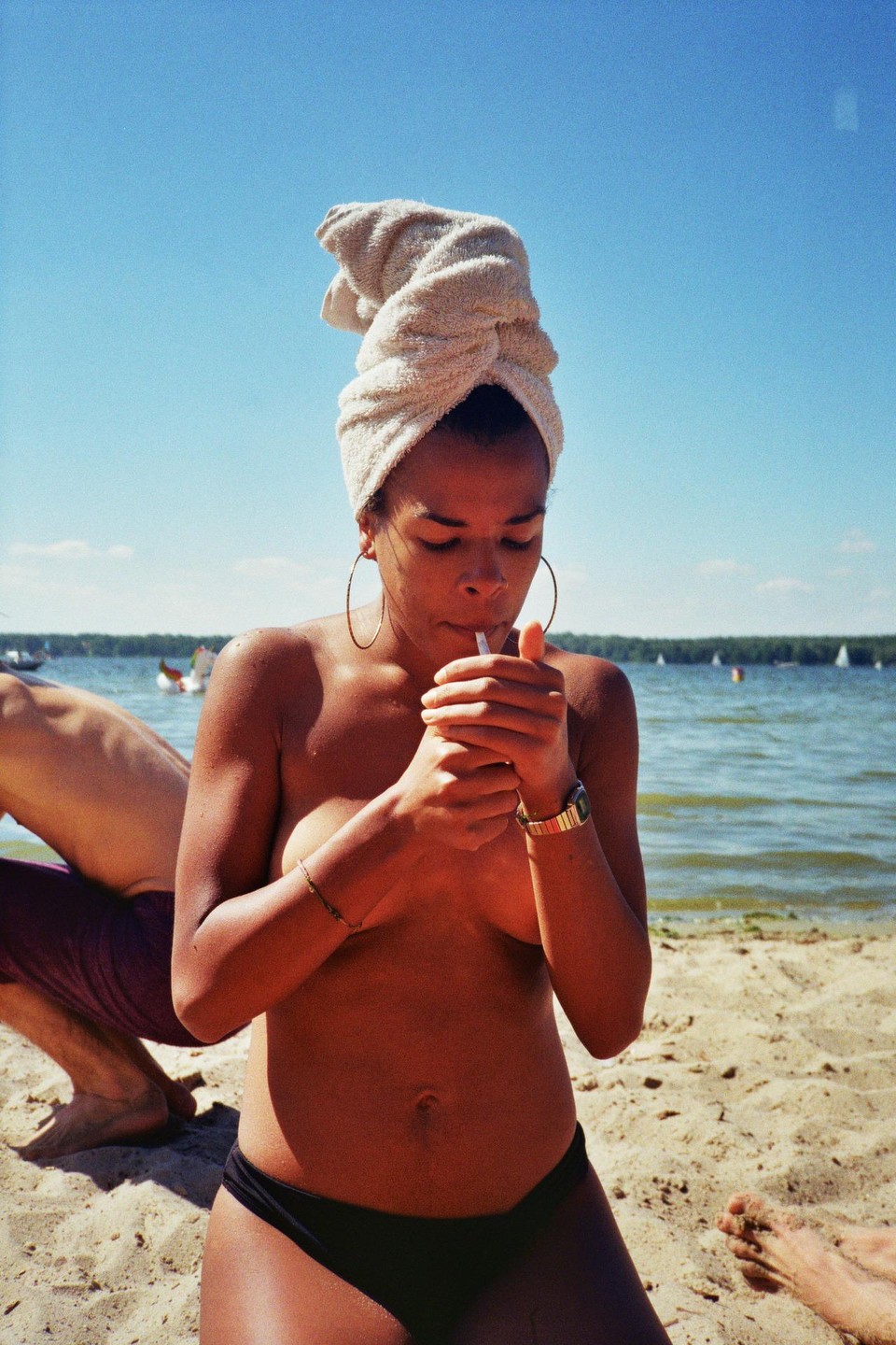A woman with a towel wrapped around her head lights a cigarette on a sandy beach next to a lake.