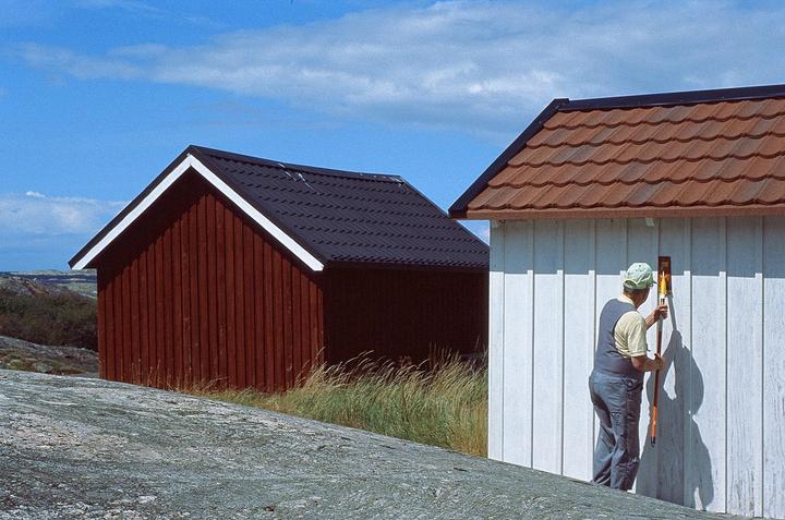 A man in a cap and overalls uses a tool to work on the side of a white building with a red roof, next to a red building with a black roof, under a blue sky.