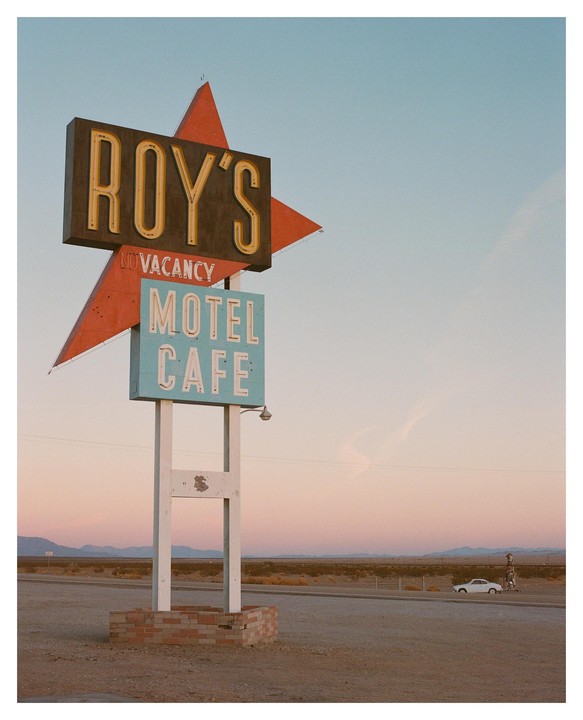 A vintage Roy's Motel and Cafe sign stands in a desert landscape with a white car parked nearby under a pastel sky.
