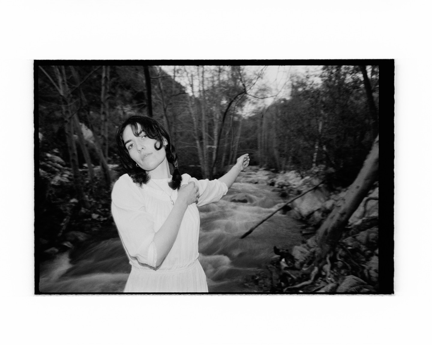 A woman in a white dress stands next to a flowing river in a forest, pointing to the right.