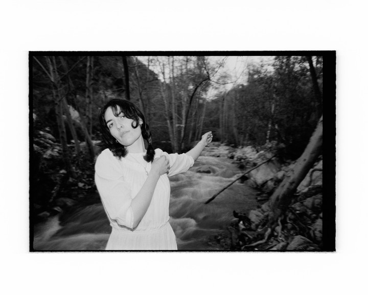 A woman in a white dress stands next to a flowing river in a forest, pointing to the right.