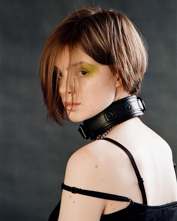 A woman with short hair and green eyeshadow looks over her shoulder while wearing a black collar.