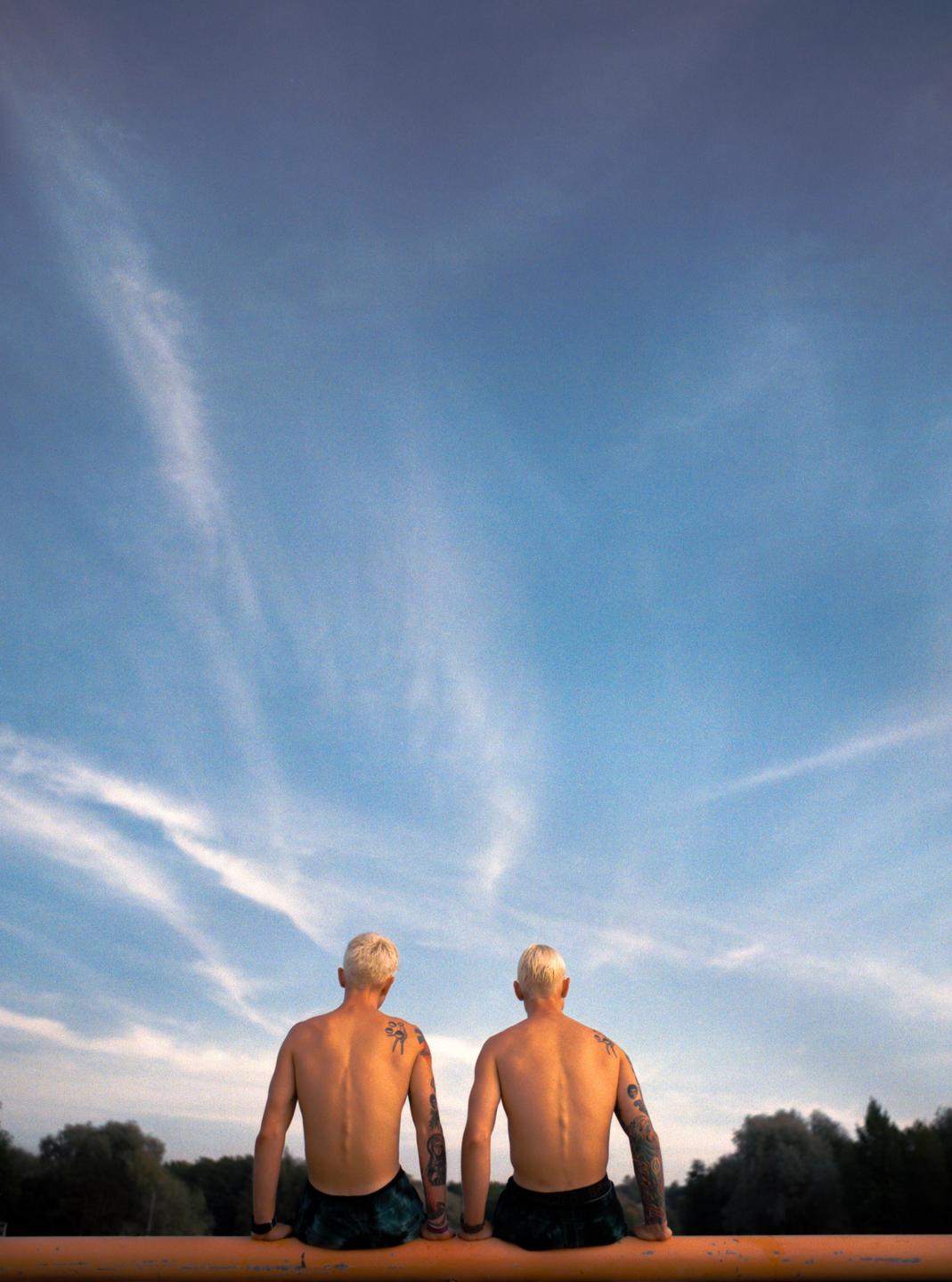 Two men with bleached hair and tattoos sit with their backs to the viewer, looking at the sky.