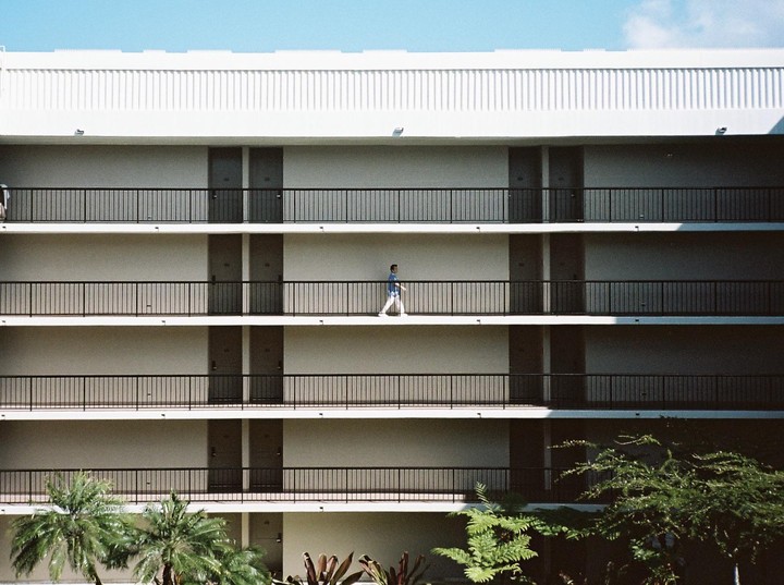 A man walks along a balcony of a multi-story building with palm trees in the foreground.