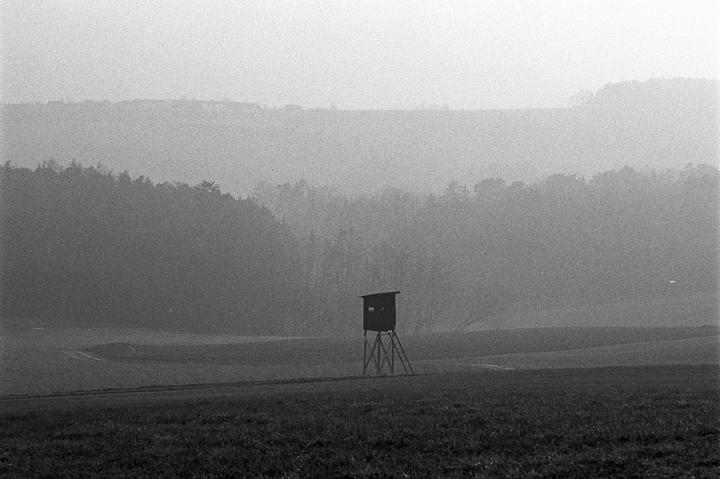 A hunting blind stands in a field with a forest and hills in the hazy background.