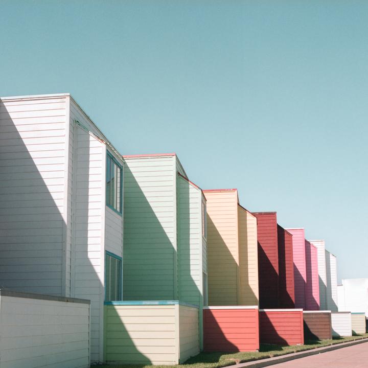 A row of colorful beach houses with strong shadows cast across their facades stands under a clear sky.