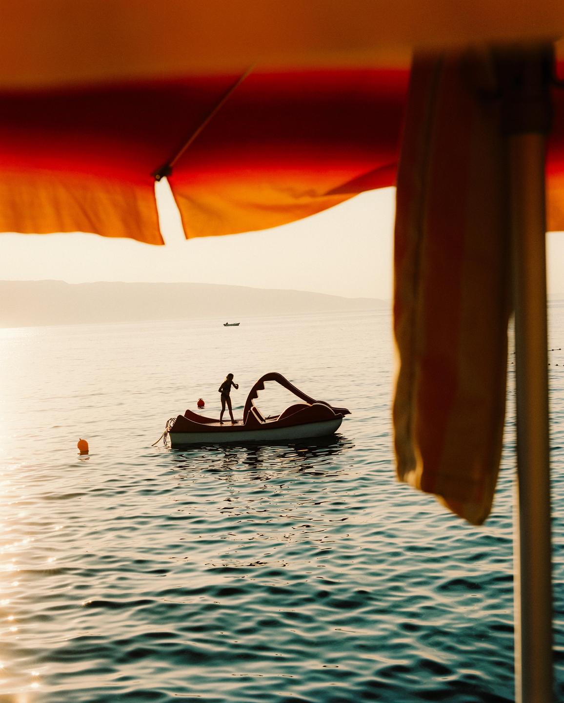 A child stands on a paddle boat in the sea, framed by a red and orange beach umbrella.