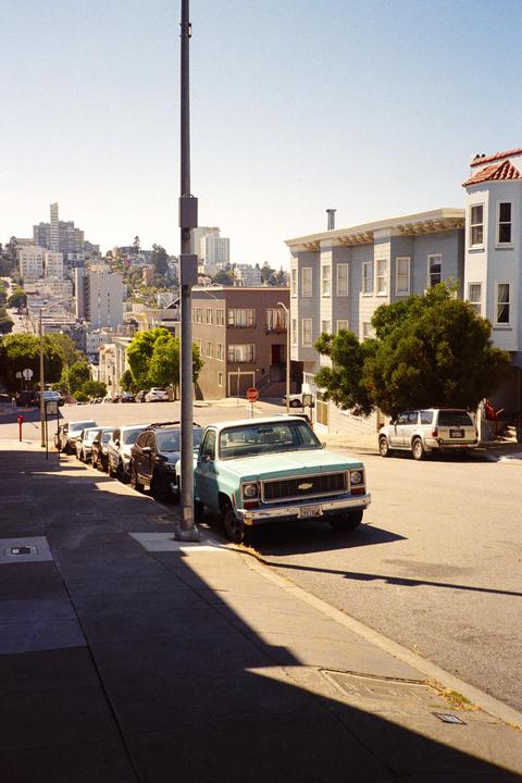 A light blue pickup truck is parked on a sloped street with other cars and buildings in the background.