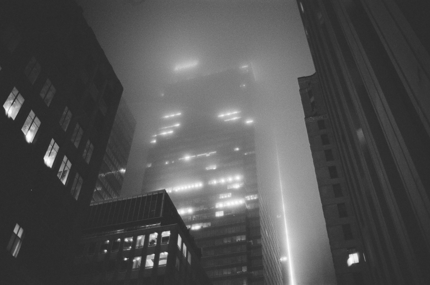 Tall buildings with illuminated windows are shrouded in fog at night.