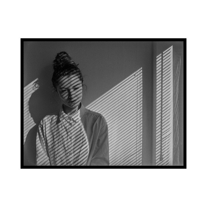A woman with her hair in a bun stands against a wall with venetian blind shadows cast across her face and body.