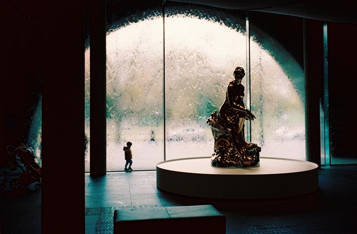 A child stands in front of a large window with water flowing down it, next to a statue of a woman.