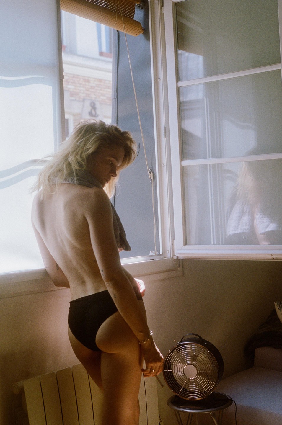 A woman stands topless in black underwear by a window with a fan on the floor.
