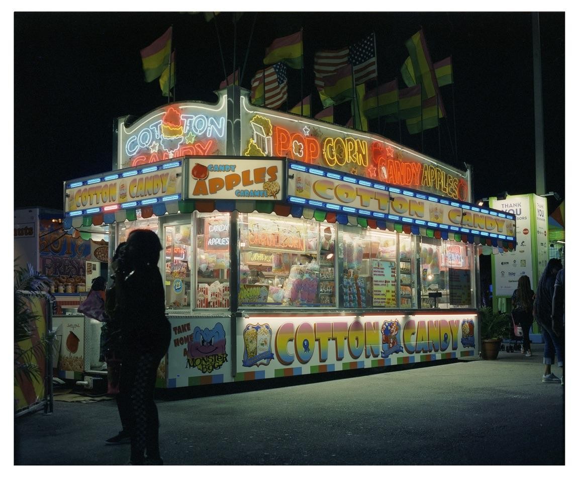 A brightly lit cotton candy and popcorn stand with neon signs stands out against the dark night sky, with flags flying above it.