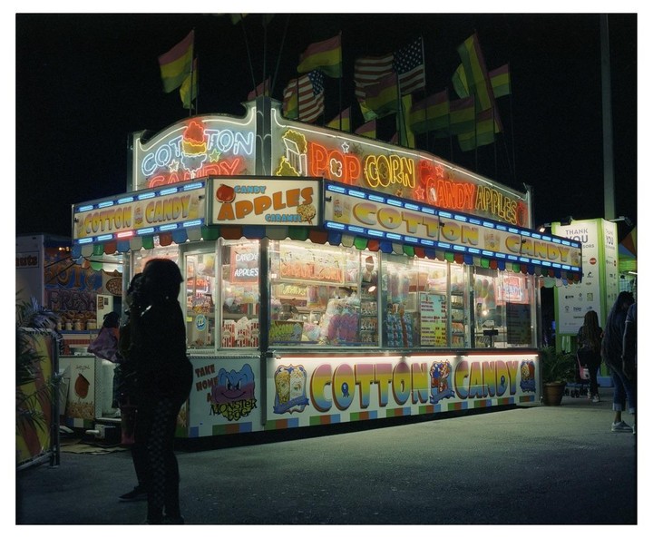 A brightly lit cotton candy and popcorn stand with neon signs stands out against the dark night sky, with flags flying above it.