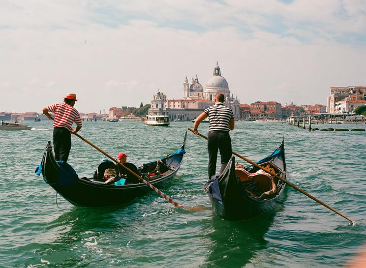 Two gondoliers stand in their gondolas on the water with a large domed building in the background.
