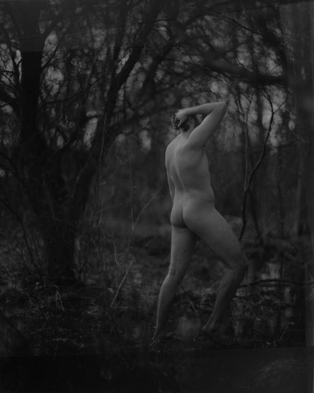 A nude man stands in a forest with his back to the viewer, his arm raised to his head.