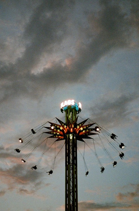 A tall swing ride with people on it is silhouetted against a cloudy sky at dusk.
