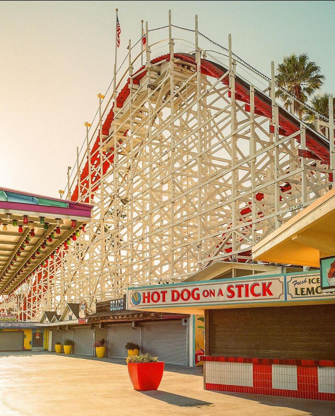 A large wooden roller coaster stands over closed storefronts on a boardwalk.