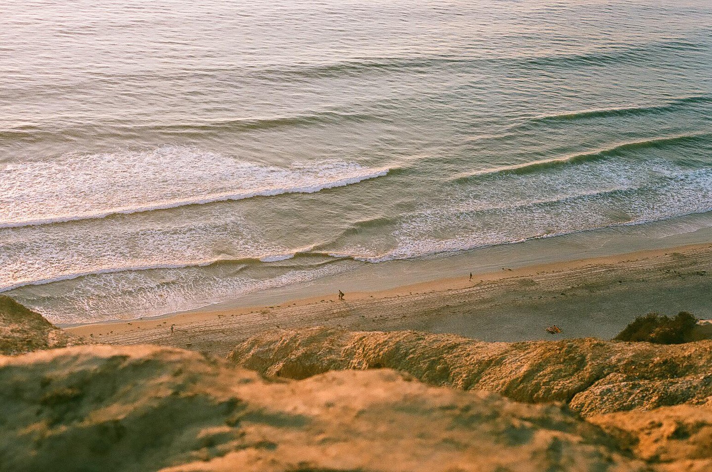 A high-angle view shows waves breaking on a beach with a few people walking on the sand.