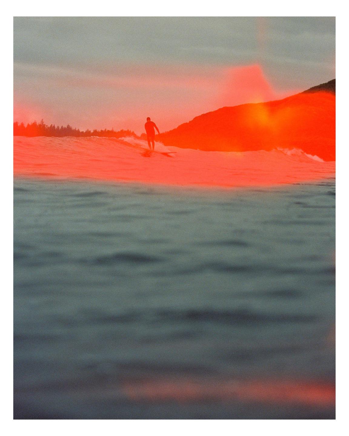 A surfer rides a wave with a bright orange light leak across the water and distant shore.