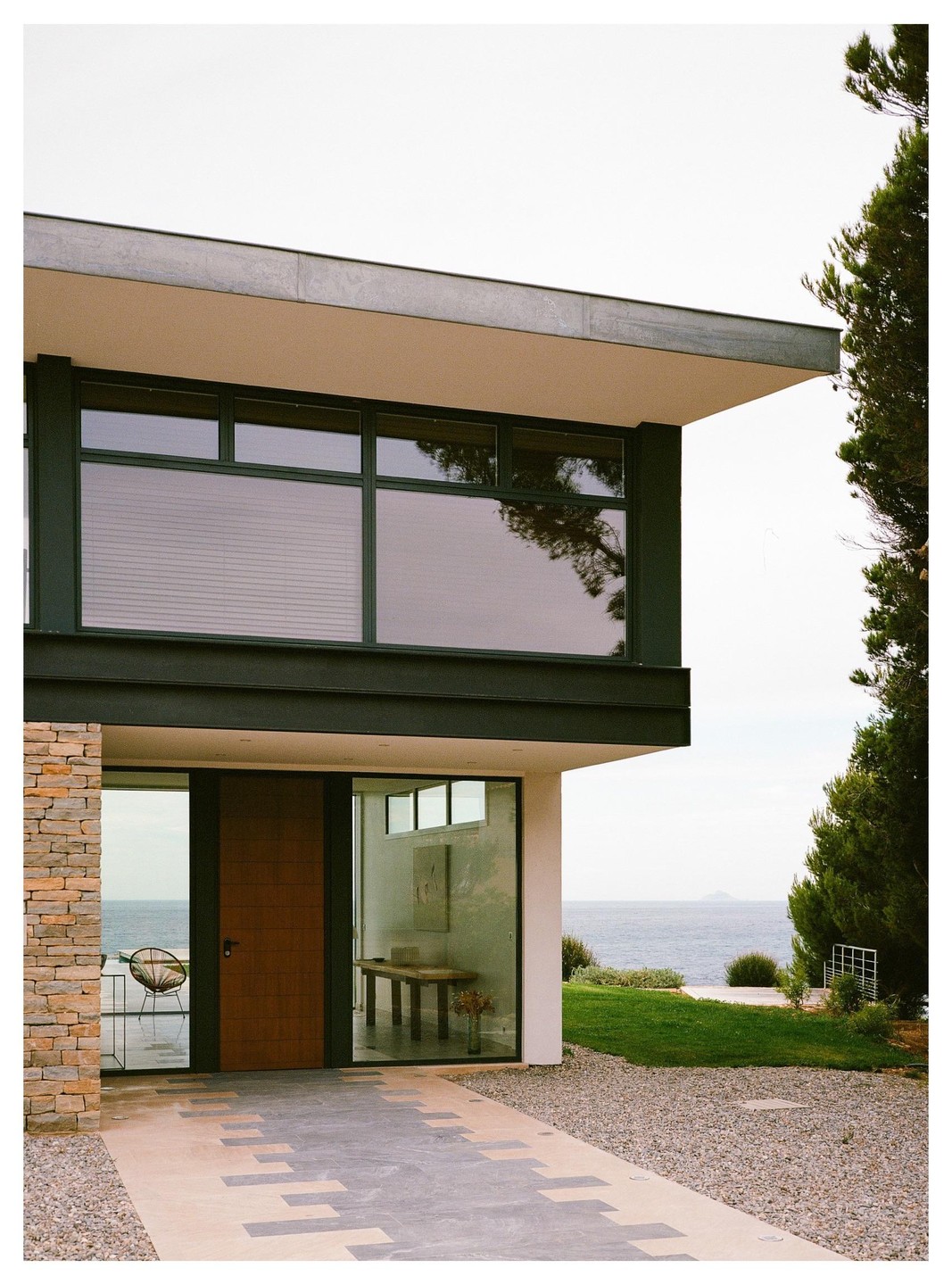 A modern house with large windows and a wooden door overlooks the ocean, with a gravel path leading to the entrance.