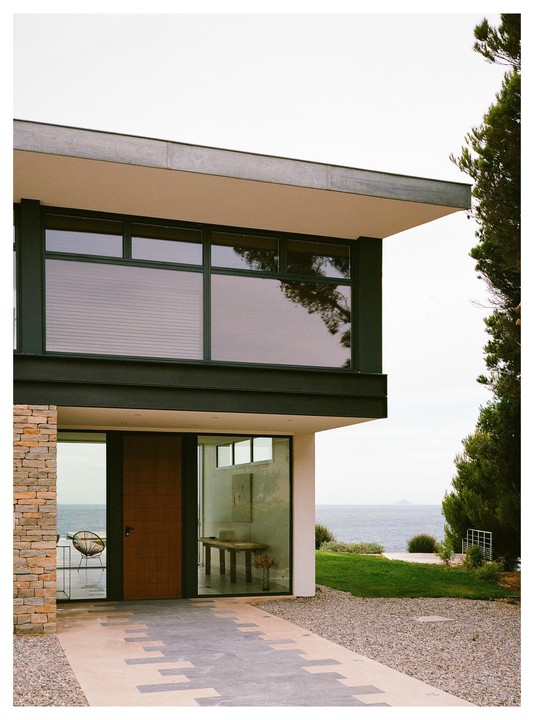 A modern house with large windows and a wooden door overlooks the ocean, with a gravel path leading to the entrance.