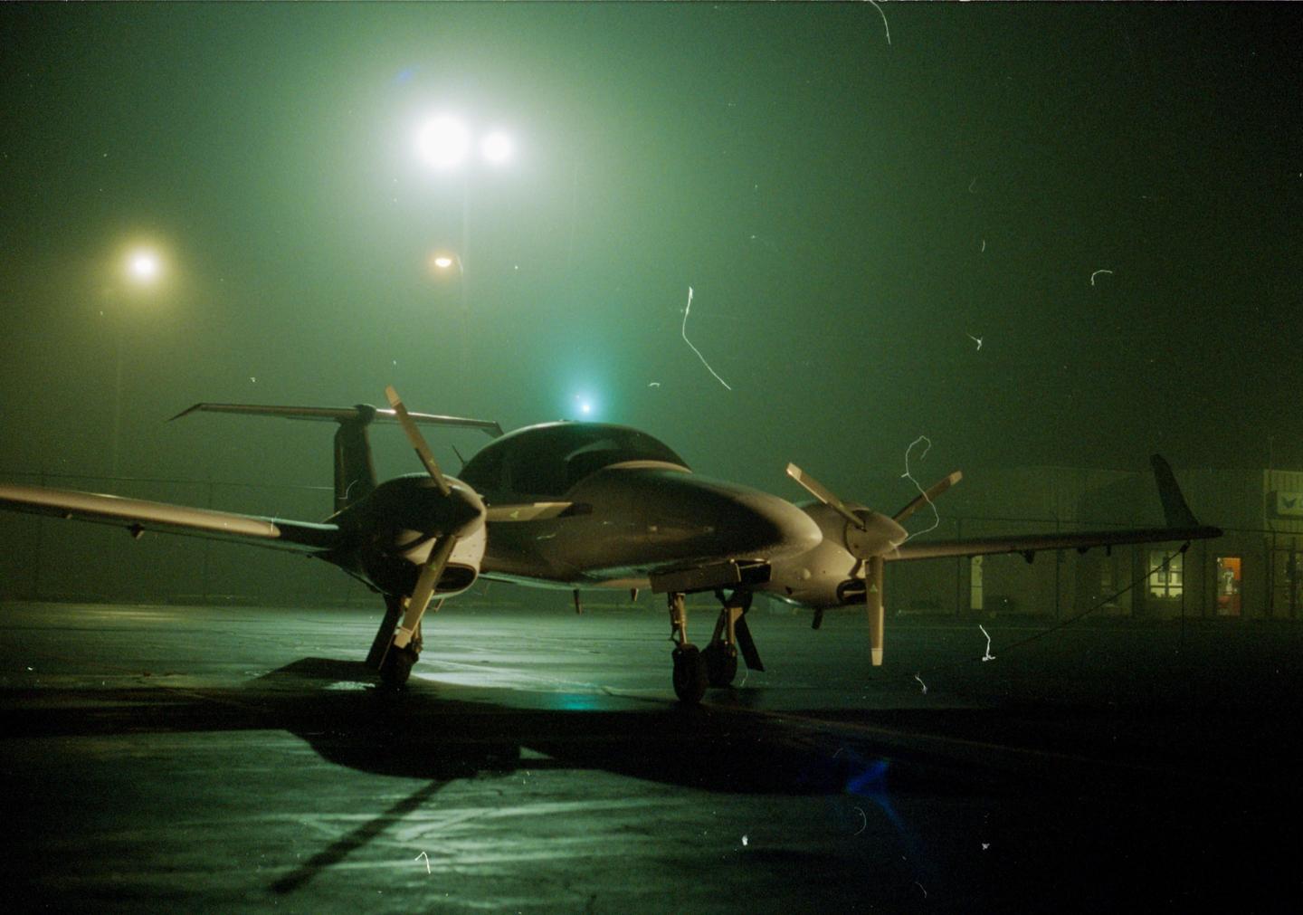 A small twin-engine propeller plane sits on an airfield at night, illuminated by bright lights.