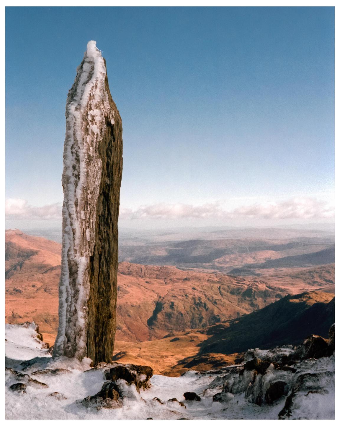 A tall, ice-covered stone pillar stands in a snowy landscape with mountains and a valley in the background under a clear sky.