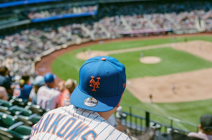 A person wearing a blue baseball cap and jersey watches a baseball game from the stands.