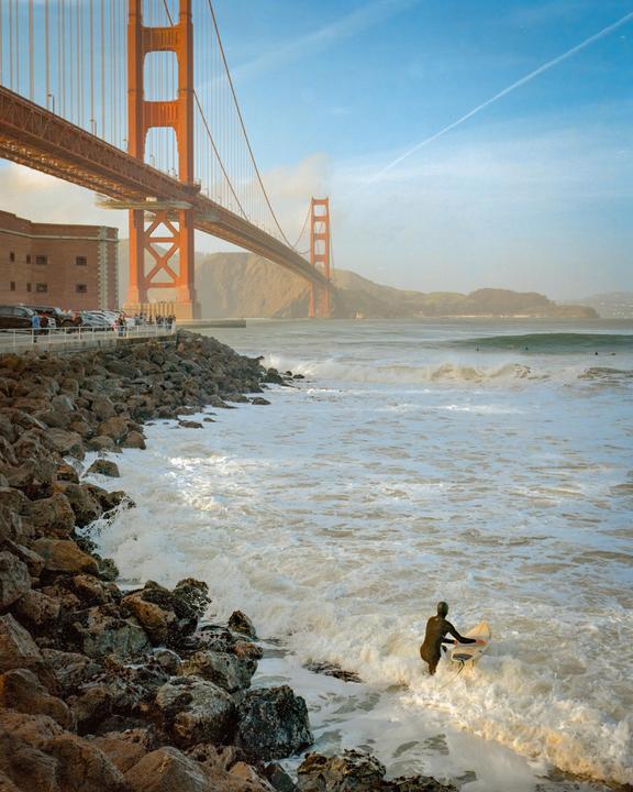 A surfer enters the ocean from a rocky shore with the Golden Gate Bridge in the background.