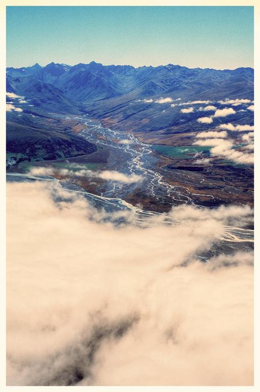 An aerial view shows a river winding through a valley surrounded by mountains, with clouds in the foreground.