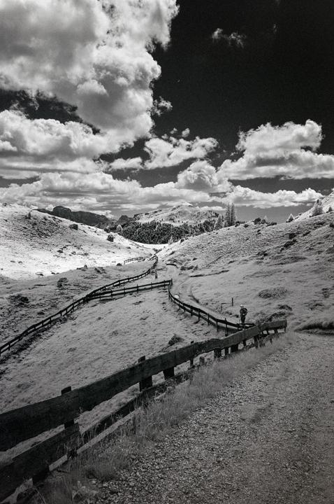 A person walks along a winding fence in a mountainous landscape under a cloudy sky.