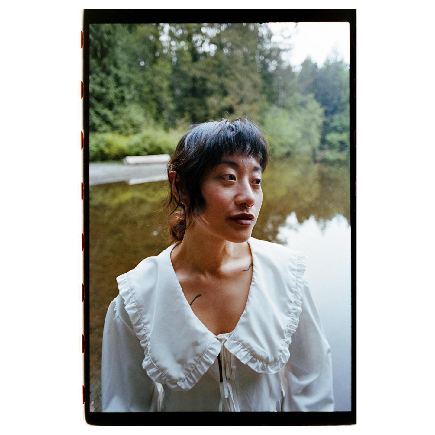 A woman with dark hair and a white ruffled collar shirt looks to the side, standing in front of a body of water and trees.