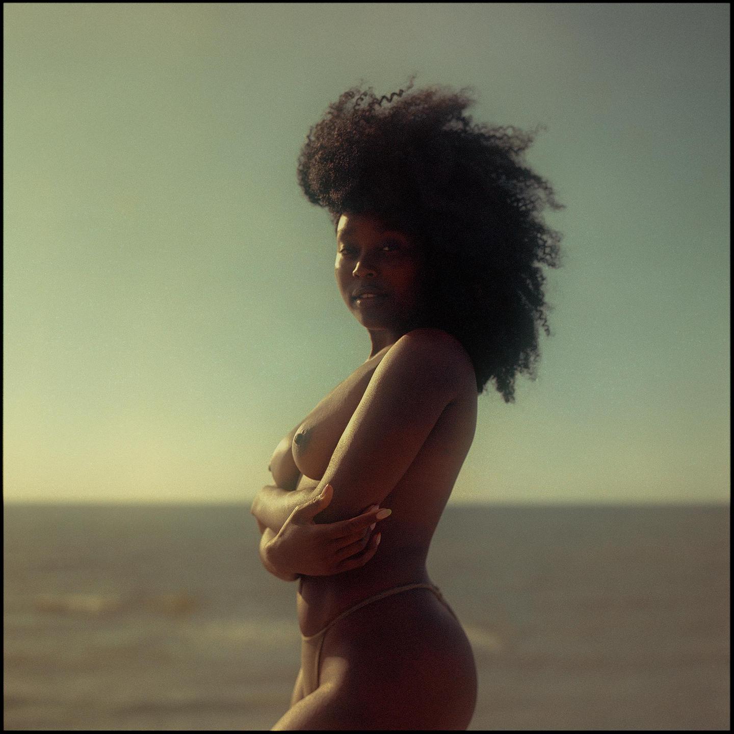 A woman with curly hair stands topless with her arms crossed in front of a body of water.